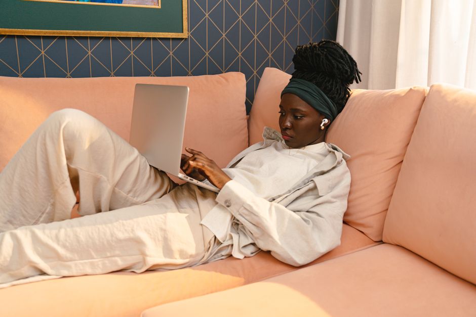 A black woman using a laptop while lounging on a cozy peach couch indoors.