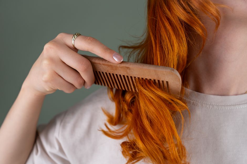 Close-up of a woman combing her red curly hair using a wooden comb against a green background.