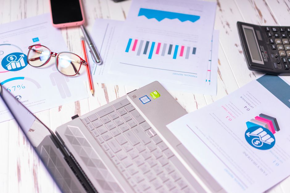 A cluttered desk with a laptop, financial reports, calculator, and glasses indicating a bu