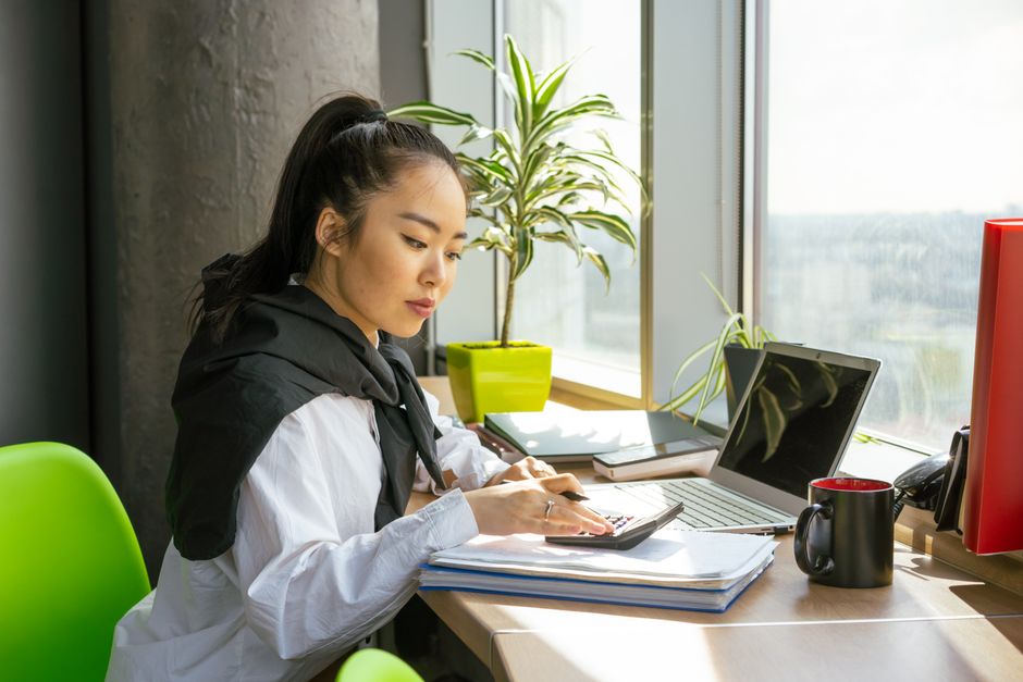 Asian woman working on finances with a calculator and laptop by a bright window in a moder