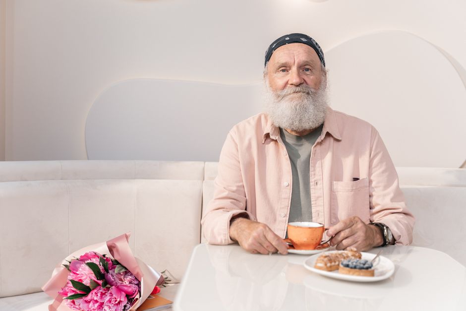 Elderly man with beard at a cafe table with coffee and pastries, bouquet of flowers beside him.