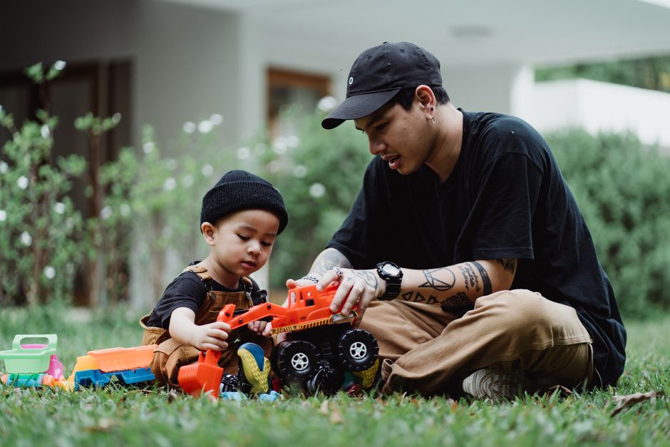 A father and son enjoy playing with toy cars on lush green grass.