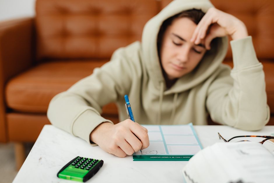 Teenage boy in a hoodie studying at home, visibly tired with a hand on his head.