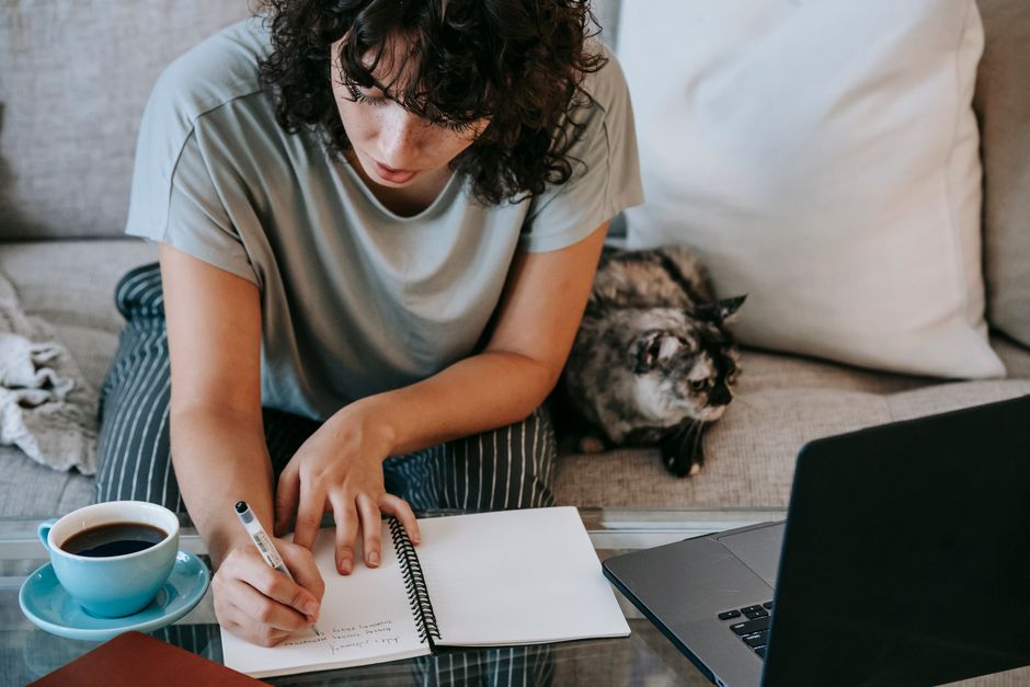 From above of crop concentrated young female student taking noted in copybook while doing homework assignment at table with laptop and cup of coffee sitting on sofa near cute cat