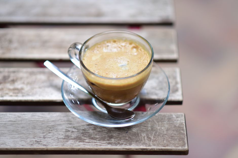 A glass cup of espresso coffee on a rustic wooden table with a spoon.