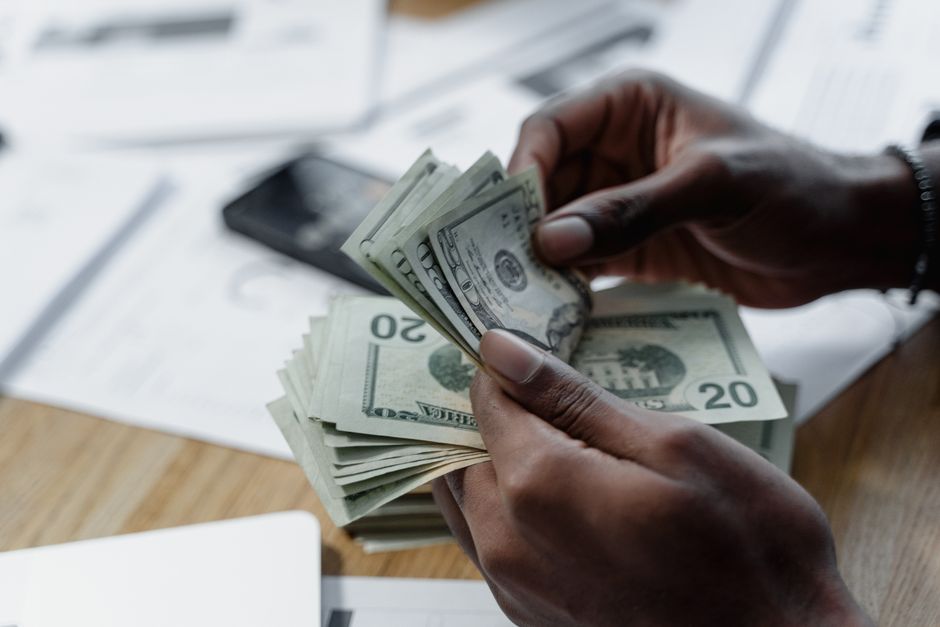 A close-up of hands counting US dollar bills on a wooden table with documents and a smartphone nearby.