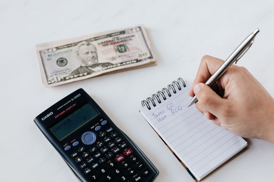 Top view of crop faceless accountant using pen and notepad while counting expenses on table near pile of paper money and calculator