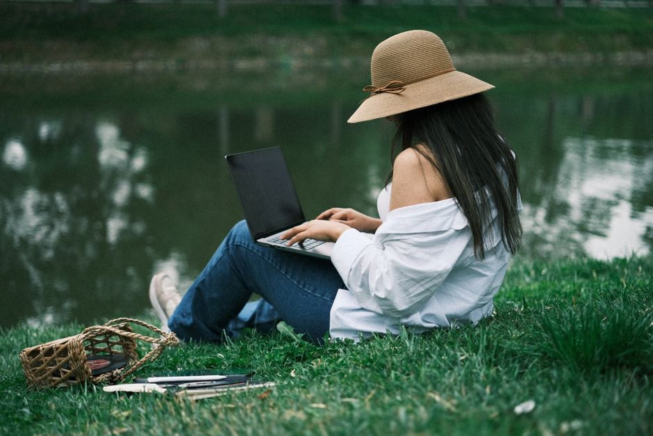 A woman in a hat sits by a riverside using a laptop, enjoying the summer day.