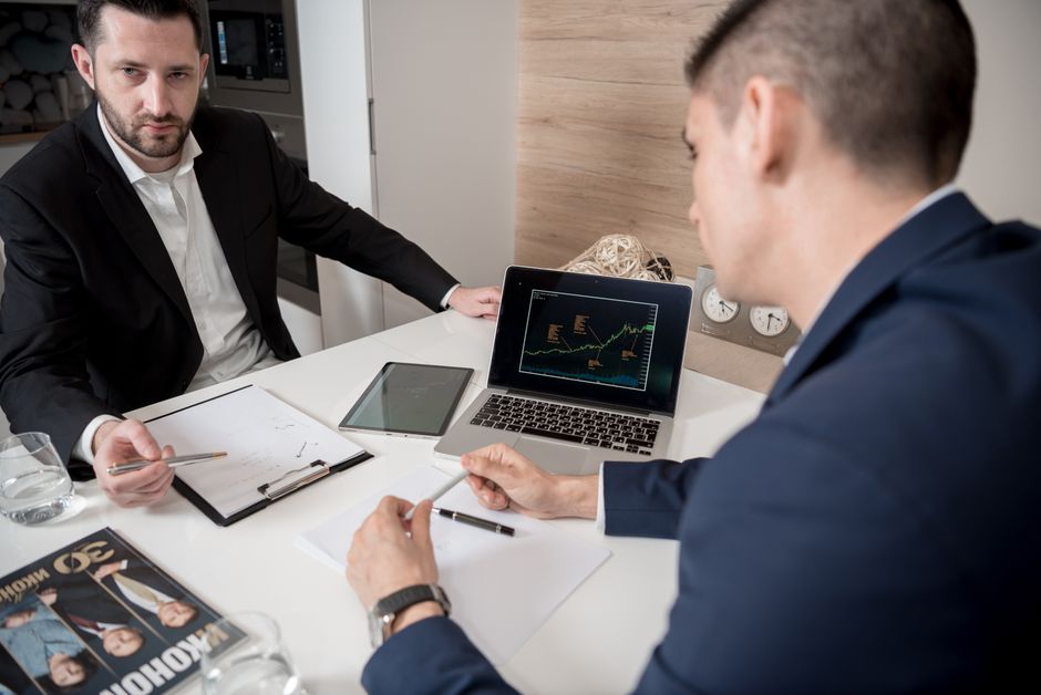 Two businessmen discussing financial graphs on a laptop and tablet in an office.